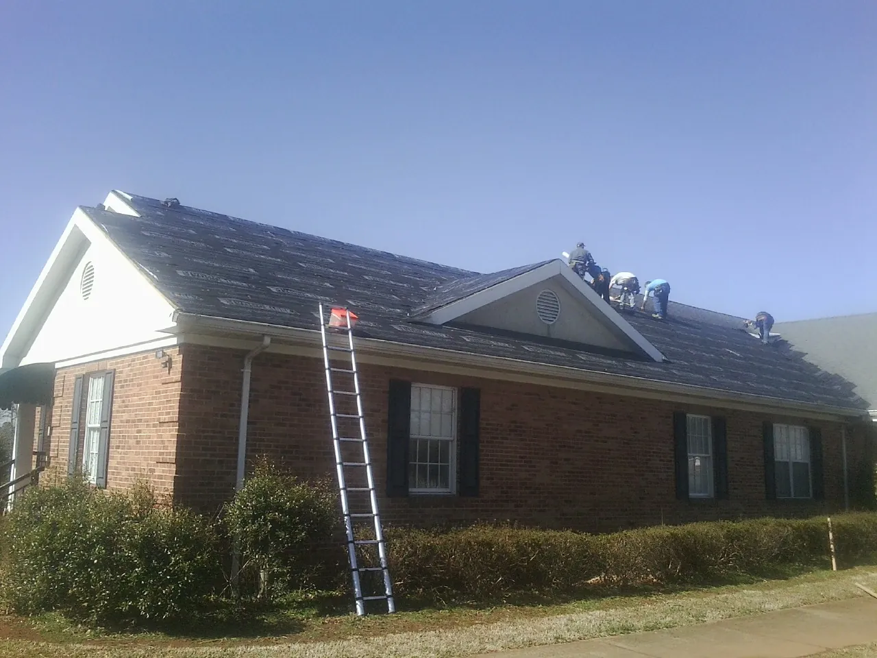 Crew tearing shingles off a brick ranch roof with a ladder set — Emergency Roof Repair work by our crew in Vinegar Bend