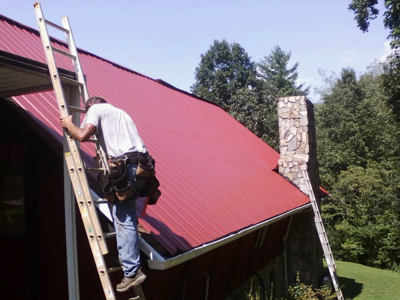 Roofer on a ladder beside a red metal roof and stone chimney in Vinegar Bend